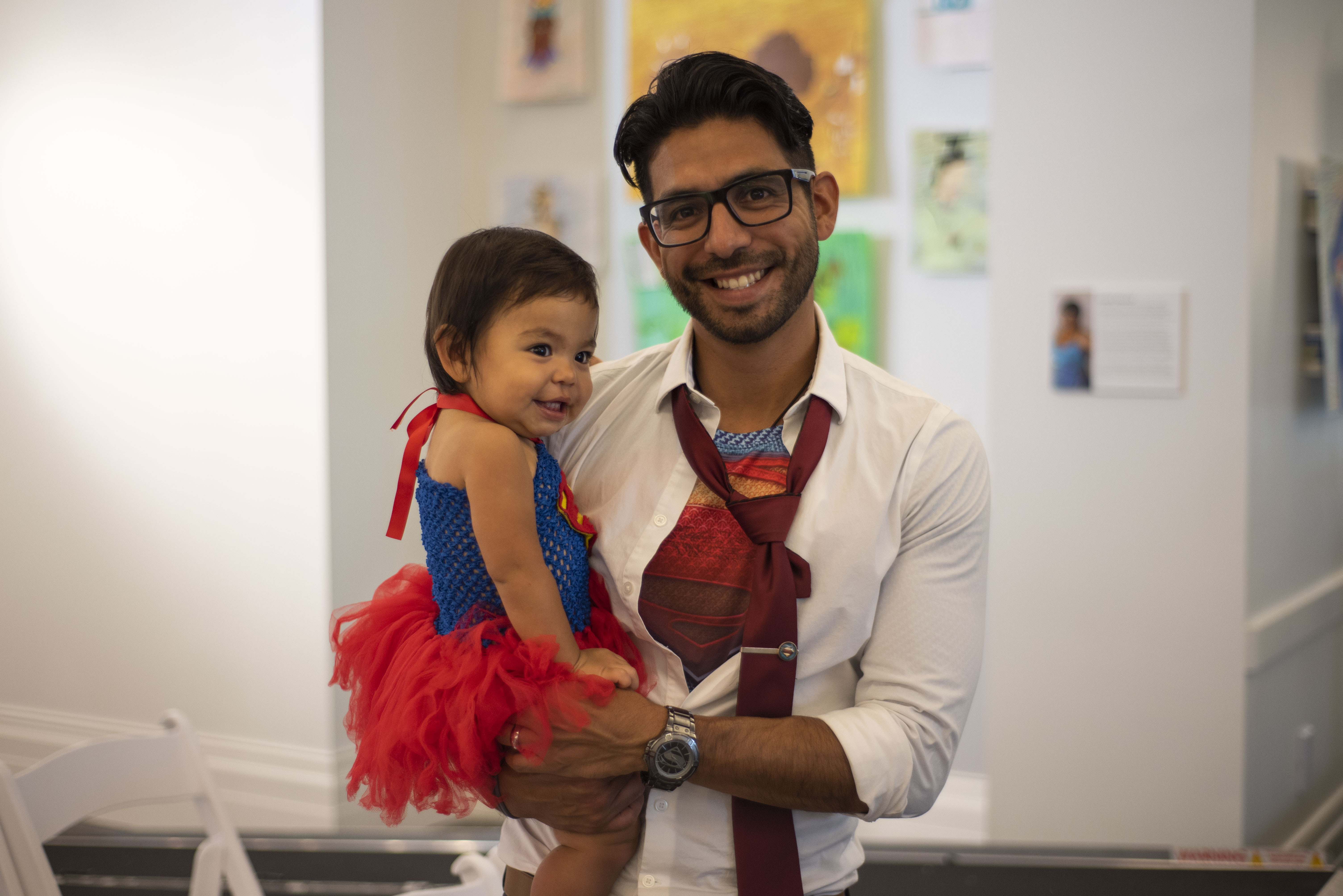 A father cosplays as Clark Kent/Superman. He's holding his daughter, who is dressed as Supergirl.