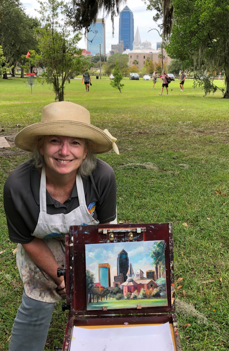 Abby Howard Murphy sits behind her plein air painting of a Jacksonville skyline