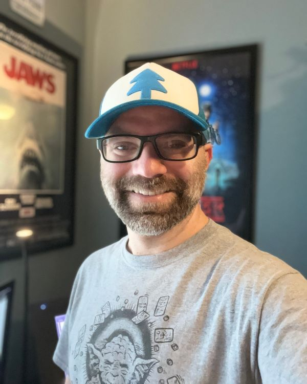 Michael Regina smiles while wearing glasses and Dipper's hat from Gravity Falls. A Jaws movie poster is behind him. 