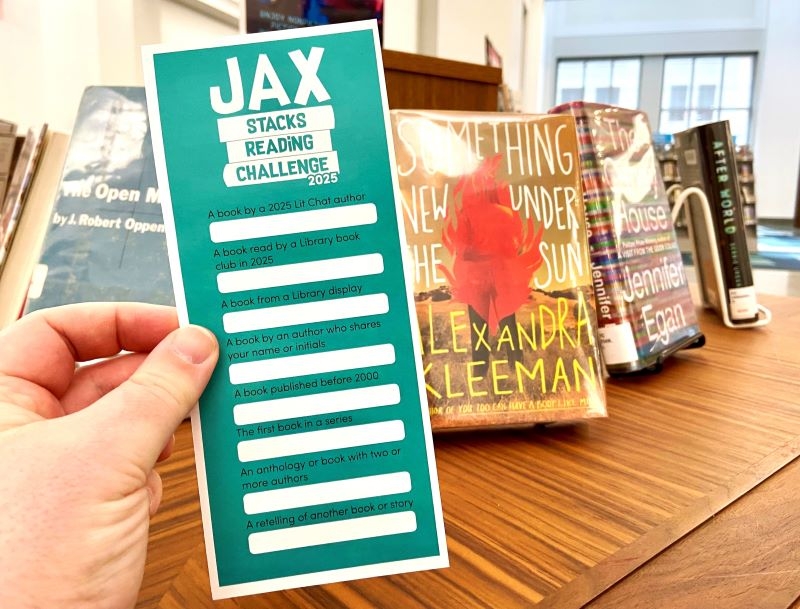 Photo of someone holding a Jax Stacks bookmark in front of a display of library books