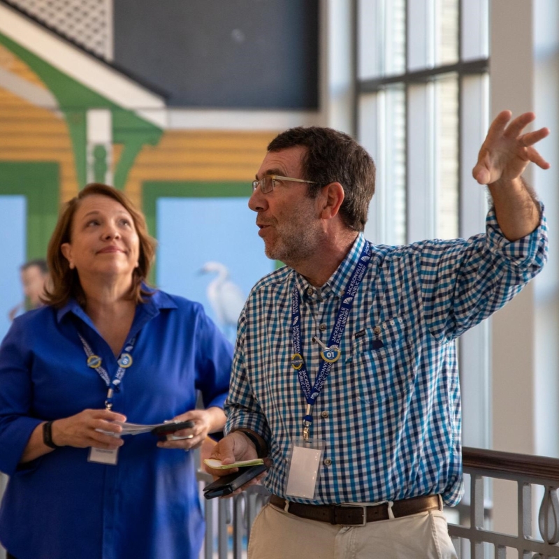 Docent gesturing to a piece of art out of frame, a mural behind him