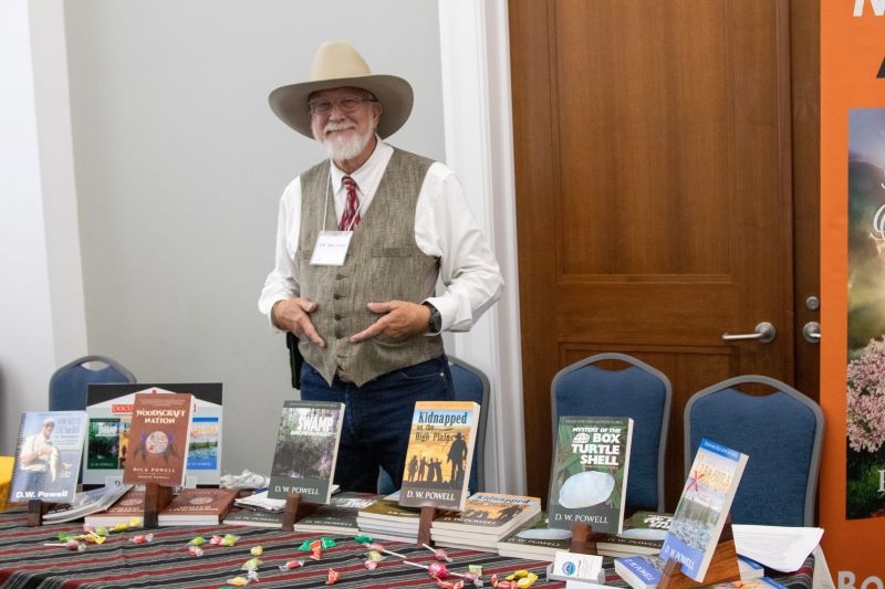 An bearded author points to a display of his books on the table in front of him.