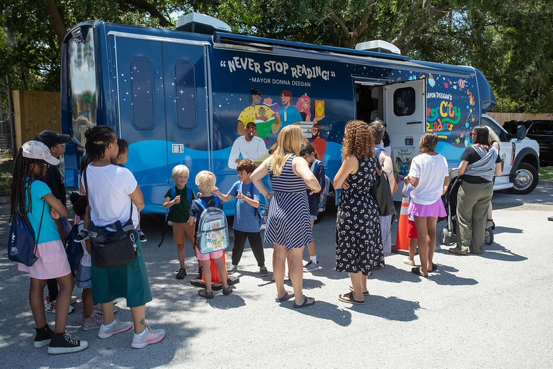 Customers waiting in line to board the bookmobile at a recent stop. The bookmobile is blue with a River City Readers logo. 
