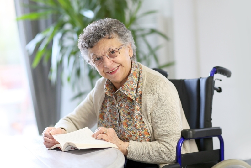 A woman using a wheelchair smiles while holding a book open on a small table