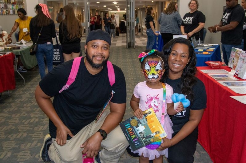A family poses for a photo in the hallway of the Conference Center. The daughter holds a book. Her face is painted like a tiger.
