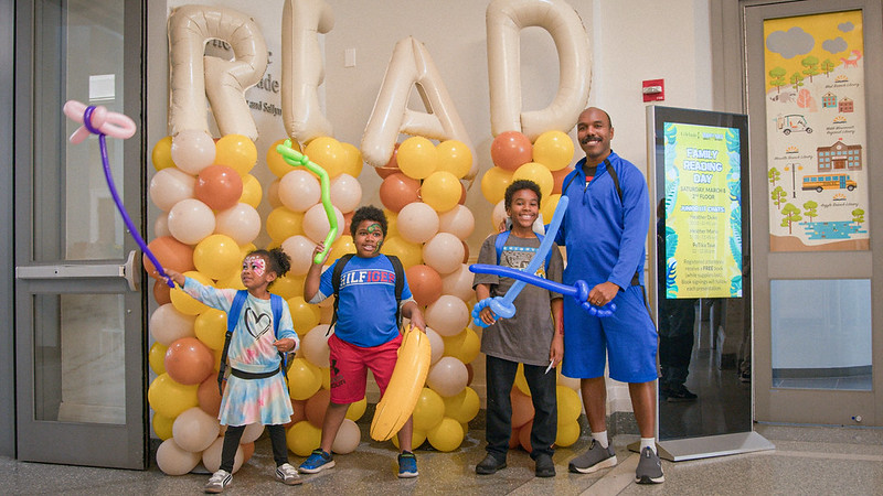 A family poses with painted faces and balloon swords in front of balloons spelling out the word 'Read.'