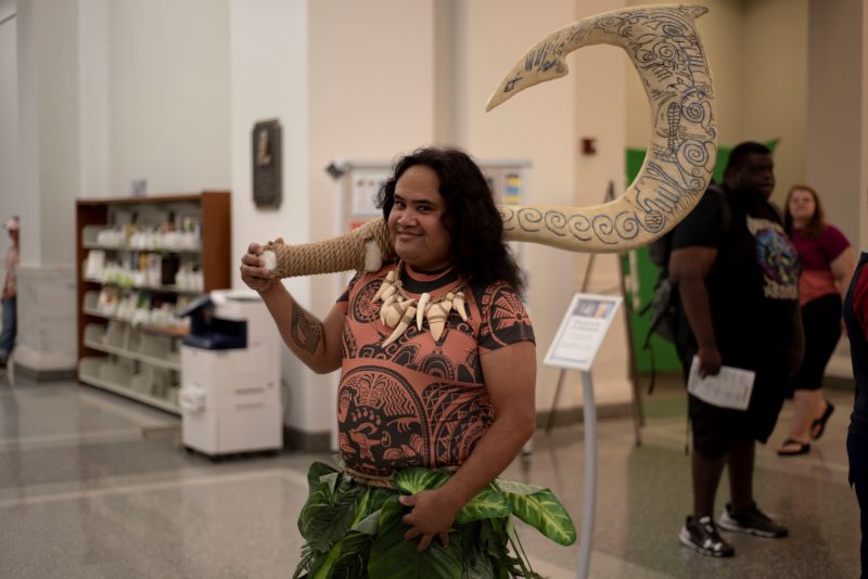 Cosplayer dressed as Maui from Moana. He's holding the character's signature hook.