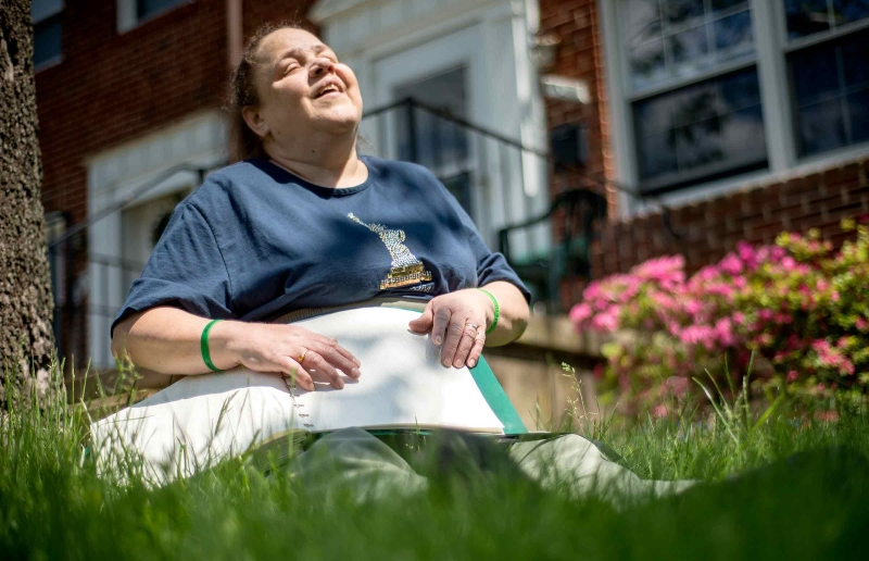 A woman reads a Braille book while sitting in the grass in front of her home