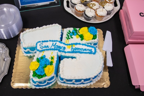 Photo of a cake in the shape of a seven and a five. It reads "San Marco 75th Anniversary." There are blue and yellow roses on top, made out of icing.