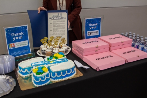 Photo of a cake, cupcakes and pink boxes of cookies on a table. A woman stands in the back holding up a Proclamation from the Mayor.