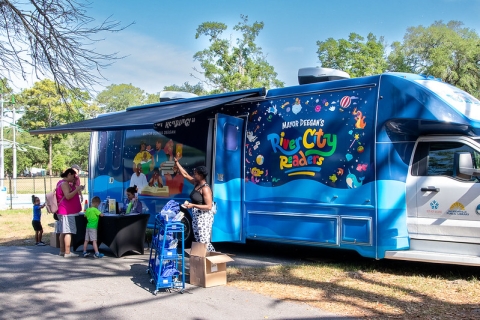 The bookmobile set up at a park. An awning extends over the side, with a small table set up underneath