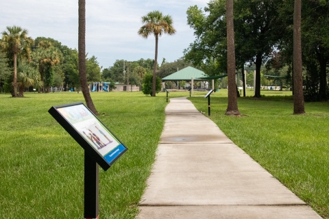 A sidewalk in the park. You can see multiple mounted signs along the path.