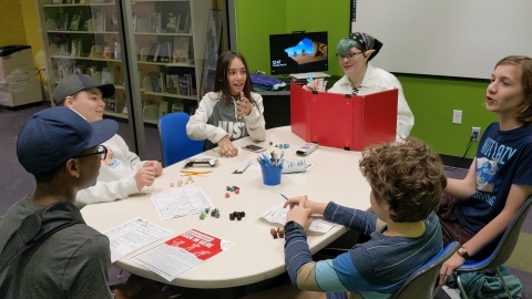 A teen Dungeon Master sits behind a DM screen at the head of the table, with teen players sitting around the table