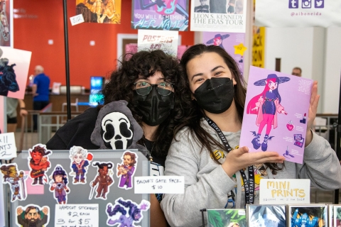 Photo of two vendors. One is holding up a small art print.