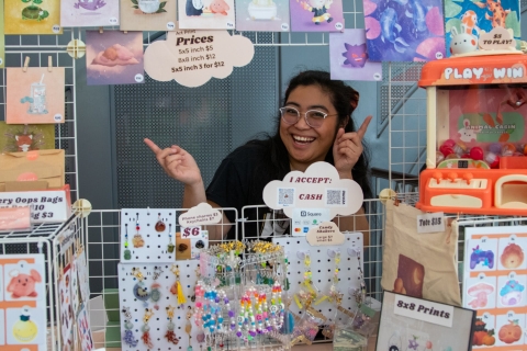 Photo of a vendor pointing to her merchandise, which includes prints, keychains and more