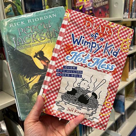 A hand holding 2 books: Percy Jackson and the Olympians and Diary of a Wimpy Kid Hot Mess.
