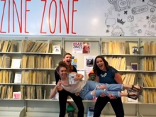 Jenna and Hurley hold Aysha Miskin in front of the Zine Zone at the Main lIbrary