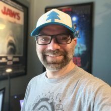 Michael Regina smiles while wearing glasses and Dipper's hat from Gravity Falls. Behind him is a Jaws movie poster. 