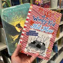 A hand holding 2 books: Percy Jackson and the Olympians and Diary of a Wimpy Kid Hot Mess.
